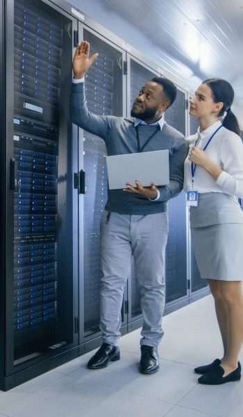 Black IT Technician with a Laptop Computer Gives a Tour to a Young Intern. They Talk in Data Center while Walking Next to Server Racks. Running Diagnostics or Doing Maintenance Work.